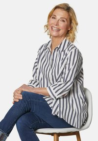 Gestreiftes Hemd in Weiß und Marineblau, mit Kragen und Dreiviertelärmeln, kombiniert mit blauen Jeans, sitzend auf einem Stuhl.