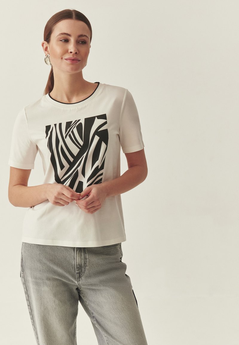 T-shirt blanc à manches courtes avec un motif abstrait noir audacieux sur le devant. Associé à un jean gris clair, ajusté et décontracté.