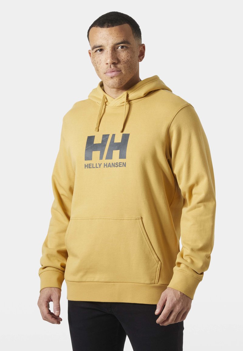 Hombre con cabello corto y pecas, vistiendo una sudadera con capucha Helly Hansen de color mostaza y pantalones negros, sobre un fondo liso.