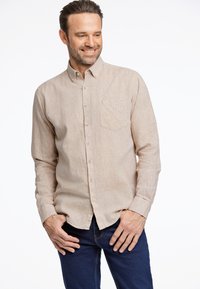 Camisa abotonada de color marrón claro con un bolsillo en el pecho, textura sutil, mangas largas y dobladillo curvado, combinada con jeans de color azul oscuro.