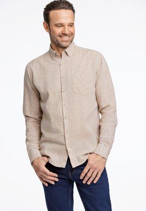 Chemise boutonnée marron clair avec une poche poitrine, texture subtile, manches longues et ourlet incurvé, assortie à un jean bleu foncé.
