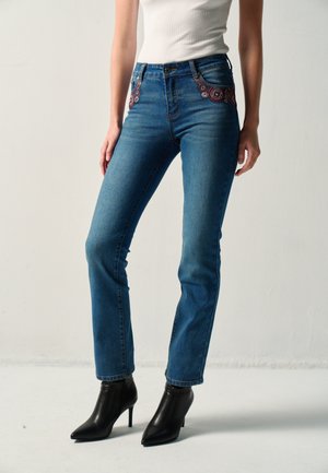 Mujer que lleva vaqueros azules de pierna recta con bordado decorativo rojo cerca de los bolsillos, top blanco sin mangas y botines negros con tacón.