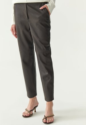 Pantalon en cuir marron avec une coupe droite, présentant des poches avant et des détails de couture, associé à des sandales à talons hauts agrémentées d'une chaîne.
