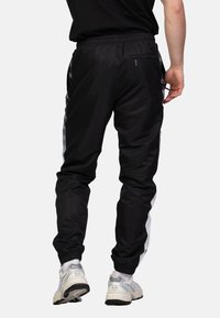 Zwarte trainingsbroek met elastische tailleband en boorden, met witte zijstrepen en een ritssluiting op de achterzak. Draagt met lichtgrijze sneakers.