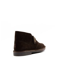 Botas de tobillo de ante marrón con puntera redonda, suela de goma plana y detalles de costura, con una etiqueta lateral que muestra el nombre de la marca "Clarks".