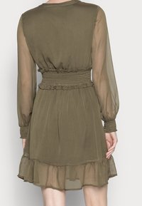 Vestido verde oliva con mangas largas transparentes, cintura fruncida y dobladillo con volantes. Textura ligera con un diseño fluido, ideal para superponer.