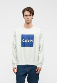 Svetlošedá mikina s modrým štvorcom, na ktorom je slovo "Calvin" tučným bielym písmom. Voľný strih s rebrovanými manžetami a pásom.