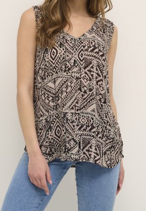 Femme portant un haut sans manches à col en V avec des motifs géométriques noirs et beiges et un jean bleu, debout devant un fond clair uni.