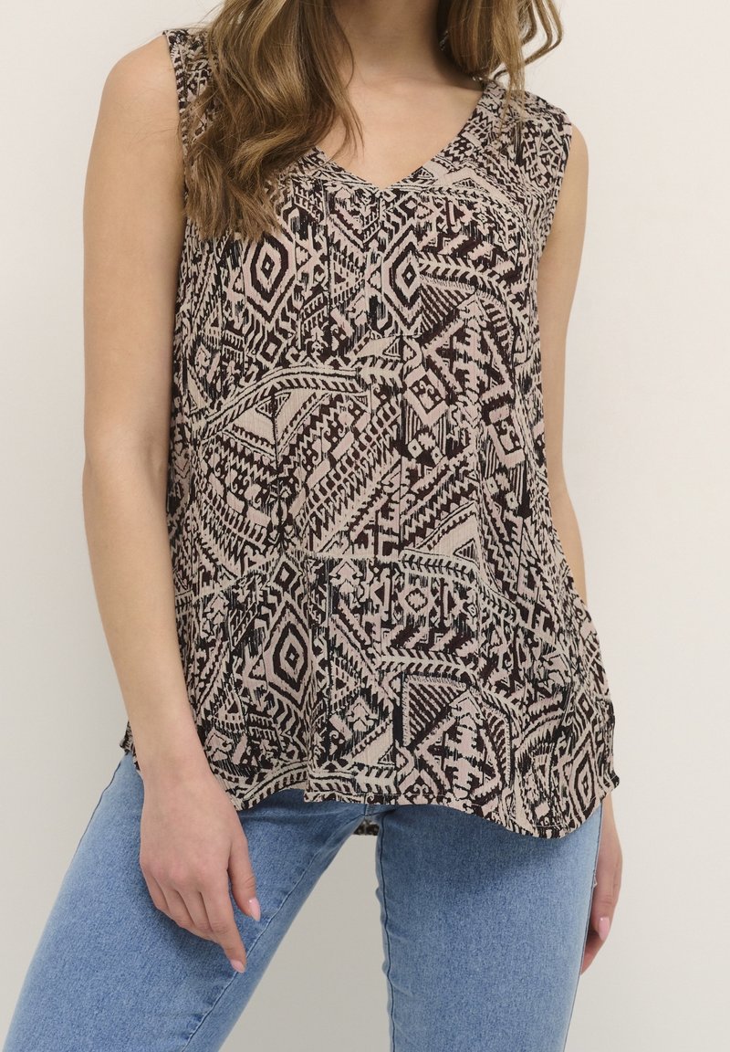 Femme portant un haut sans manches à col en V avec des motifs géométriques noirs et beiges et un jean bleu, debout devant un fond clair uni.