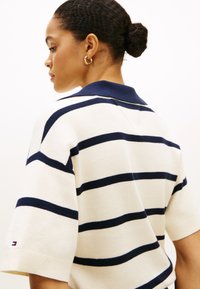 Ριγέ πόλο πουκάμισο σε κρεμ χρώμα με ναυτικές ρίγες, με κοντά μανίκια και λαιμόκοψη με γιακά, κατασκευασμένο από πλεκτό ύφασμα. Λεπτομέρεια με λογότυπο στο μανίκι.