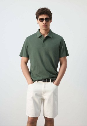 Hombre con camiseta polo verde oscuro, pantalones cortos blancos, cinturón negro y gafas de sol, de pie con las manos en los bolsillos contra un fondo liso.