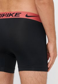 Sorte atletiske shorts med en tætsiddende pasform. Har en rød talje med sort "Nike" logo. Glat tekstur og længde til midt på låret.