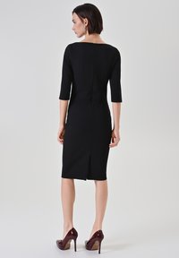 Robe noire ajustée avec manches trois-quarts, fermeture éclair au dos et fente au genou, associée à des escarpins bordeaux brillants à talons hauts.