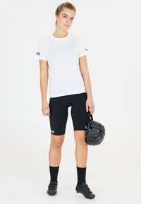 T-shirt de sport blanche, shorts de cyclisme noirs, chaussures de cyclisme noires et casque noir, debout sur un fond blanc uni.