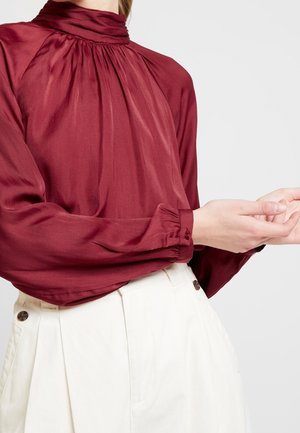 Blouse bordeaux à col montant et détails froncés, manches longues avec poignets élastiques, accompagnée de pantalons blancs taille haute ornés de boutons.