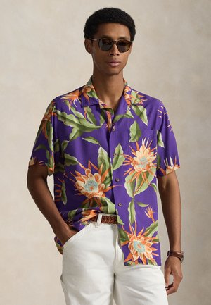 Άνδρας που φοράει σκούρα γυαλιά ηλίου και μοβ κοντομάνικο πουκάμισο με μεγάλα πορτοκαλί και πράσινα ανθρωπίδια σχέδια, συνδυασμένο με λευκό παντελόνι.