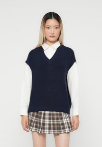 Pull en tricot sans manches bleu marine avec un col en V, porté par-dessus une chemise blanche à col. Assorti avec une jupe à carreaux aux tons marron et crème.