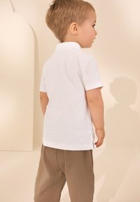 Weißes Poloshirt mit kurzen Ärmeln aus strukturiertem Stoff, kombiniert mit braunen Shorts. Die Rückansicht zeigt ein schlichtes Design und klare Linien.