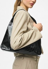 Moteris, vilkinti smėlio spalvos kostiumą, laiko didelę blizgią juodą odinę rankinę priešais vientisą foną.