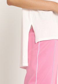 Top corto blanco de manga corta con aberturas laterales, combinado con pantalones deportivos rosas que tienen ribetes blancos a lo largo de los lados. Diseño simple, textura suave.