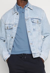 Ljust blå denimjacka med två bröstfickor, knappstängning och sömnadsdetaljer, bärs över en blå polo och mörka byxor.