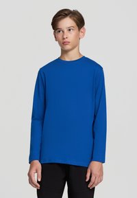 Koszulka z długim rękawem, z okrągłym dekoltem w jasnym niebieskim kolorze, o gładkiej teksturze i luźnym kroju. Noszona z czarnymi spodniami.