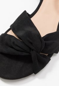 Sandale plate en suède noir avec un design de nœud en façade. Semelle lisse avec une semelle intérieure légèrement rembourrée pour le confort.