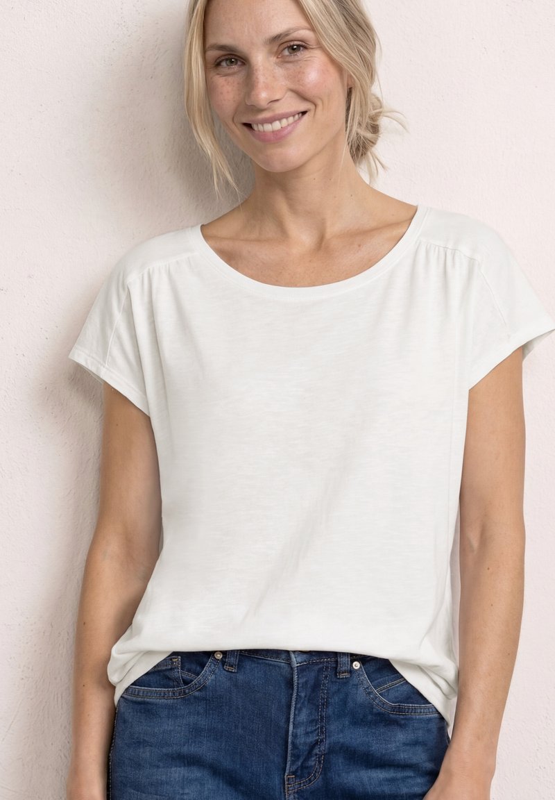 Lächelnde Frau mit blond zurückgebundenem Haar, die ein weißes Kurzarmhemd und blaue Jeans trägt und vor einer hellen Wand steht.
