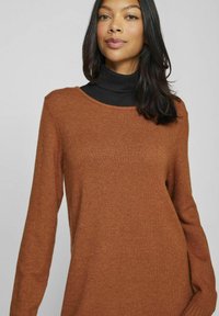 Maglione marrone a maglia con scollatura ampia e maniche lunghe. La texture sembra morbida e leggermente pelucchosa, adatta per essere indossata sopra un dolcevita nero.