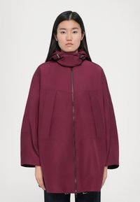 Veste à capuche bordeaux avec une fermeture éclair avant, coupe oversized, deux grandes poches et un tissu lisse au toucher mat. Design simple.