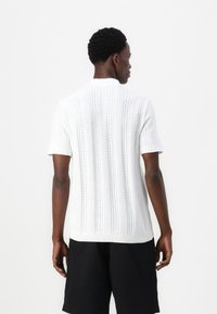 Balts īsām piedurknēm polo krekls ar adītu tekstūru, kuram ir vertikālas svītru raksts un ribota apkakle. Zem tā ir uzvilktas melnas šortus.