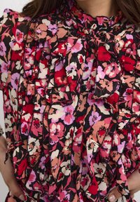 Kukallinen pusero, jossa on rypytetty muotoilu, pääasiassa punaisissa, vaaleanpunaisissa ja mustissa sävyissä, mustilla napeilla ja tekstuurilla.