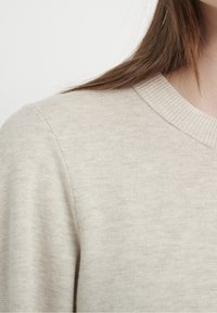 Pull beige en maille avec un col rond, une bordure côtelée et une texture douce, présentant un motif chiné subtil et un détail de couture d'épaule.