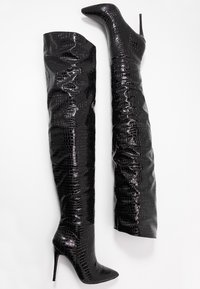 Botas negras hasta la rodilla con un acabado brillante y textura de cocodrilo estampada. Con un diseño de punta afilada y tacón de aguja. Diseño alto y ajustado.