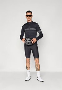 Svart sykkelklær med lange ermer og shorts, med en horisontal grå stripe. Modellen har på seg solbriller og holder en svart hjelm.