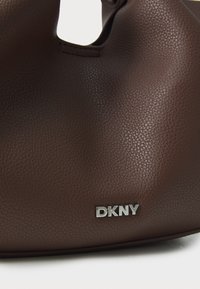 Geantă din piele maro cu textură piatră, având o formă rotunjită, dotată cu un accent argintiu cu logo-ul DKNY pe lateral. Textură netedă, fără modele vizibile.