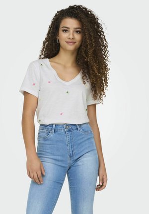 T-shirt blanc à col en V avec broderie florale colorée, manches courtes, associé à un jean taille haute bleu clair. Texture de tissu lisse.