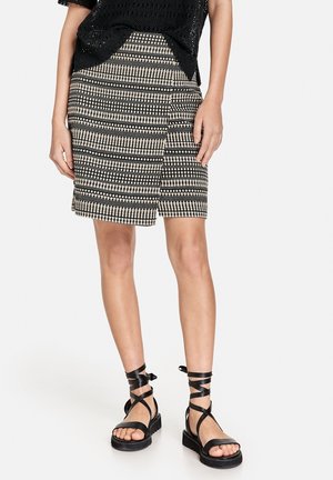 Vrouw draagt een zwarte en beige patterned wikkelrok, zwarte gebreide top en zwarte platte sandalen met enkelbandjes.