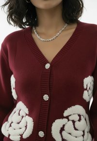 Bordowy sweter na drutach z białym kwiatowym wzorem, z dekoltem w serek, srebrnymi guzikami oraz prążkowanymi wykończeniami na krawędziach.