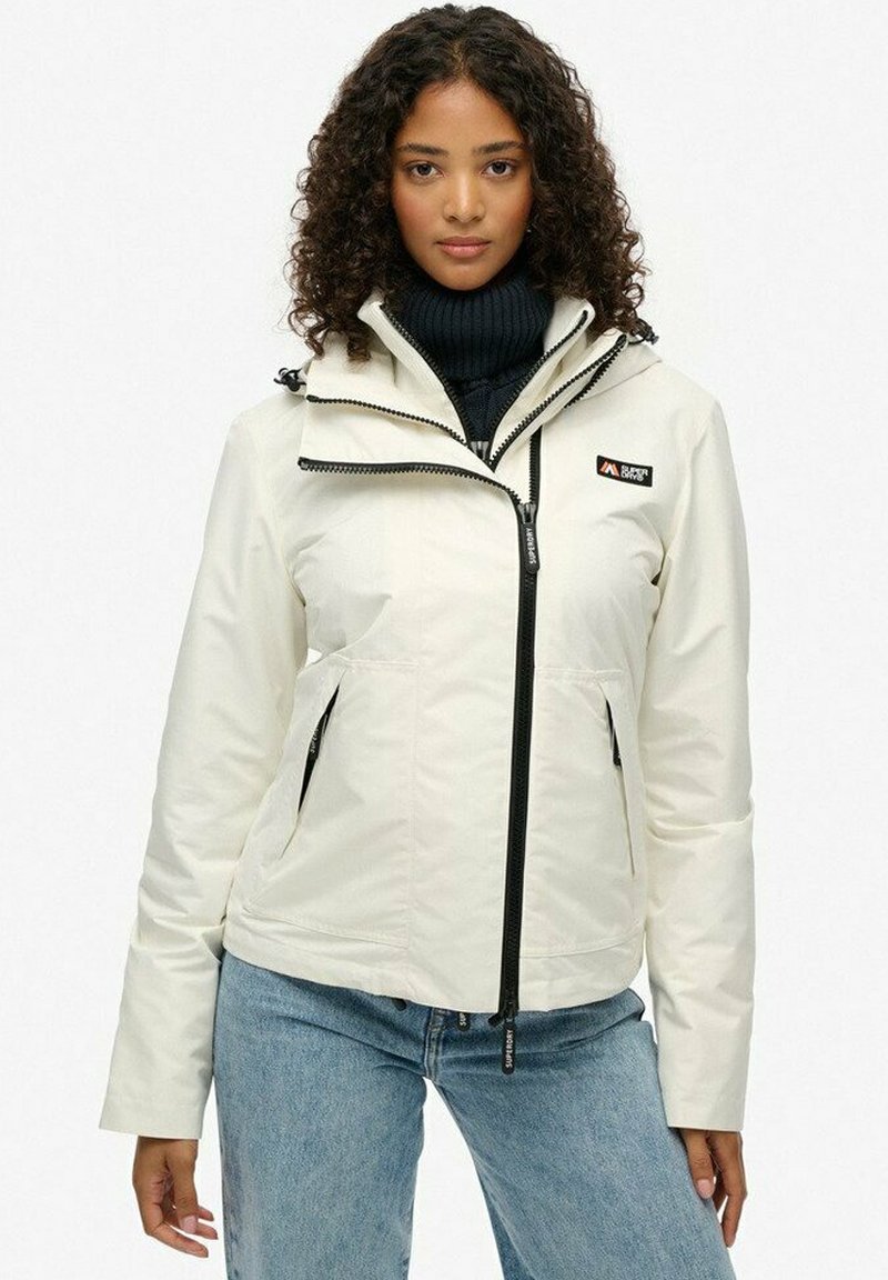 Kvinde med krøllet hår iført en hvid zip-up jakke over en sort turtleneck og blå jeans, stående foran en ensfarvet hvid baggrund.