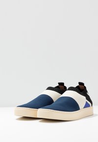 Stickade slip-on-skor i blått, krämfärgat och svart med ett strukturerat mönster, vadderad ankel och en vit gummisula.