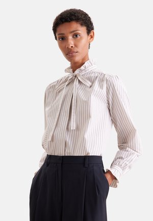 Vrouw draagt een witte blouse met lange mouwen met dunne verticale strepen en een strik in de nek, gecombineerd met zwart hooggetailleerde broek.