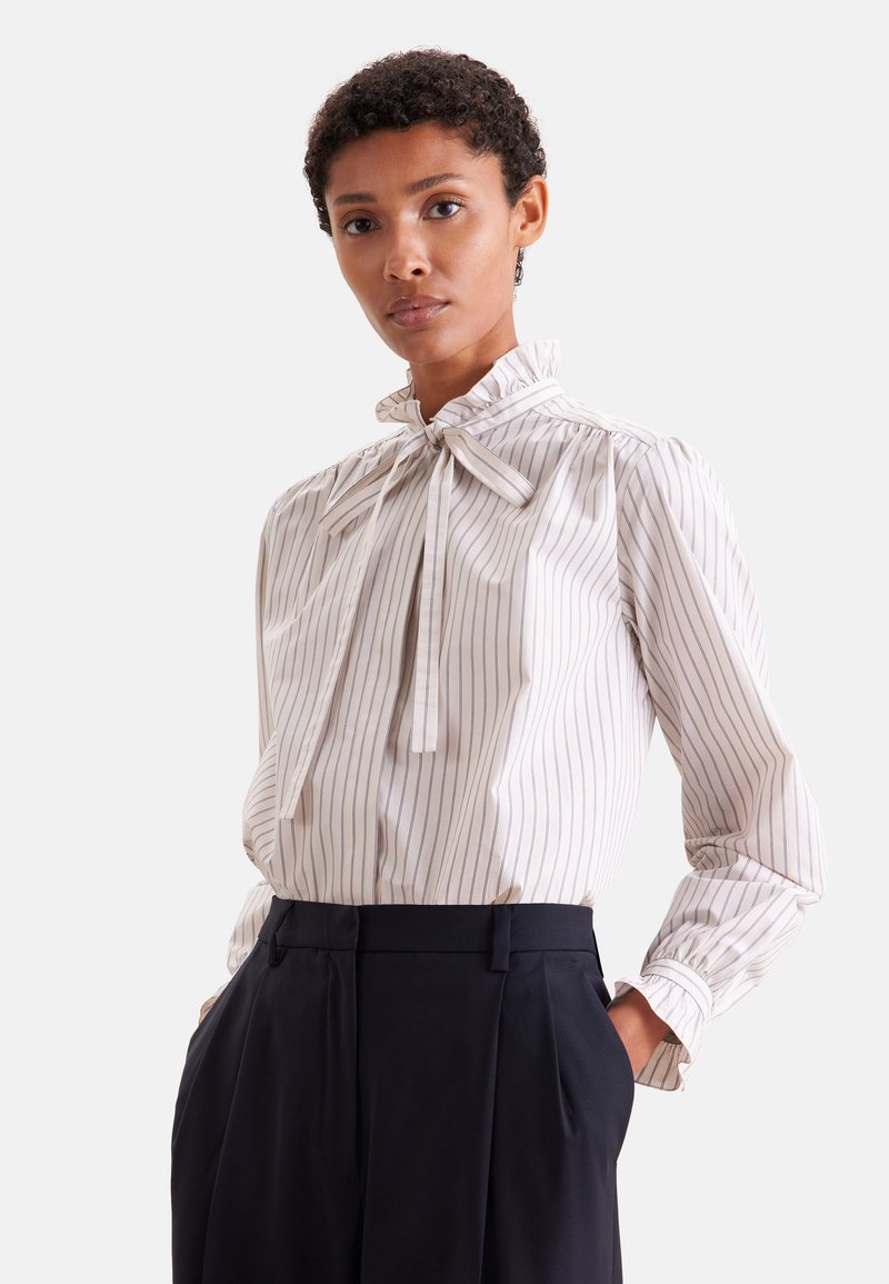 Femme portant un chemisier blanc à manches longues avec de fines rayures verticales et un nœud au cou, associé à un pantalon noir taille haute.