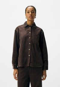 Brown corduroy shirt with a button-down front and a pocket. Features long sleeves and a textured finish, paired with matching pants.