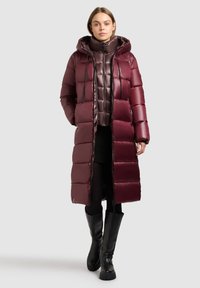 Femme portant un long manteau matelassé bordeaux avec capuche, superposé à une doudoune courte brillante, une jupe noire, des collants et des bottes noires montantes.