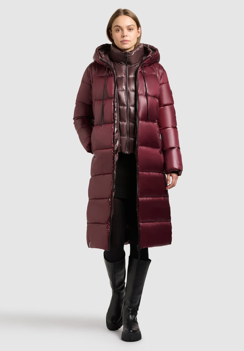 Femme portant un long manteau matelassé bordeaux avec capuche, superposé à une doudoune courte brillante, une jupe noire, des collants et des bottes noires montantes.
