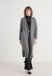 Grå strikket lang cardigan med åpen front, sammen med en svart ribbestrikket topp og svarte bukser. Har en avslappet silhuett og teksturert stoff.