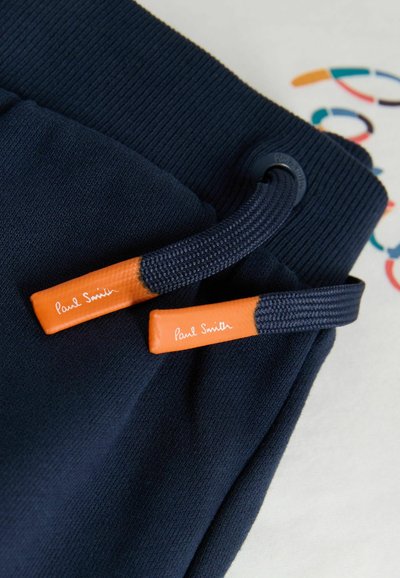 Ceinture de pantalon de survêtement bleu marine avec de larges cordons de serrage terminés par des embouts orange et le texte "Paul Smith" en blanc, posée sur un fond en tissu blanc.