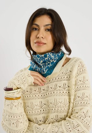 Mujer joven con suéter de crochet color crema, bufanda azul con flores y tres pulseras en la muñeca izquierda, posando frente a un fondo liso.