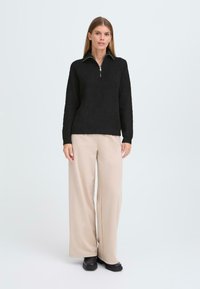 Pulover tricotat negru cu guler tip zip. Textură ondulată și mâneci lungi, asociate cu pantaloni largi bej și pantofi negri.